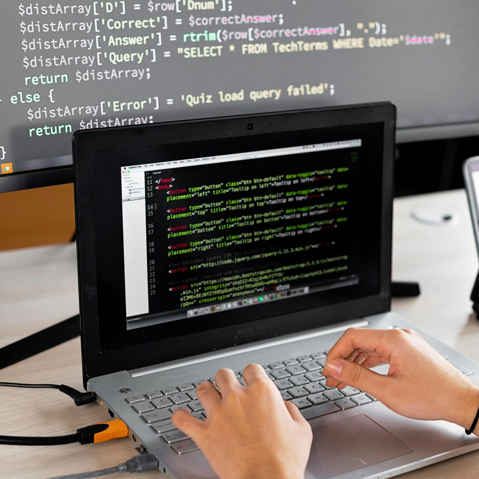 Person coding at a desk with laptop and external monitor showing programming code.